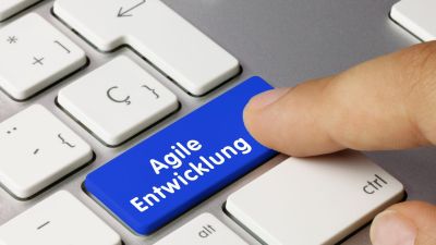 Finger drückt blaue Taste mit Aufschrift „Agile Entwicklung“ auf Computertastatur als Symbol für agiles Projektmanagement und moderne Softwareentwicklung. 