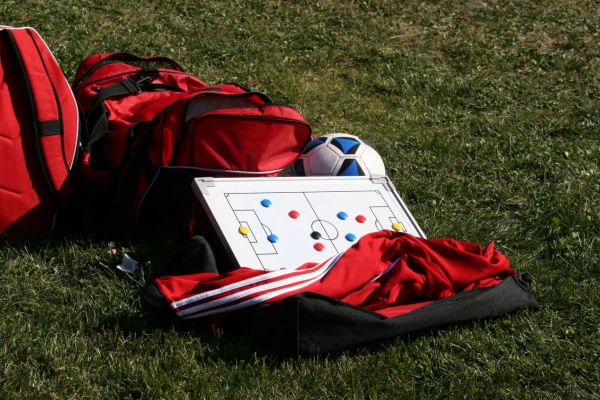 Rote Sporttaschen, Fußball und Taktiktafel mit Spielfeldmarkierung auf grünem Rasen: Symbol für Strategie, Planung und Teamarbeit im Projektmanagement.