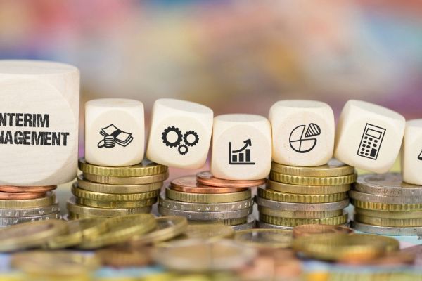 Holzwürfel mit Symbolen zu Finanzen, Prozessen und Kennzahlen stehen auf gestapelten Münzen: Visualisierung des ROI von Interim Management im Unternehmen. 