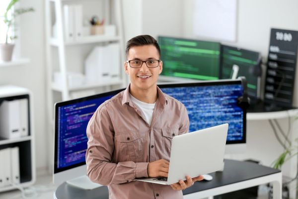 IT-Spezialist mit Laptop im modernen Büro vor Monitoren mit Programmiercode: Symbol für Personaldienstleistung und IT-Fachkräfte. 