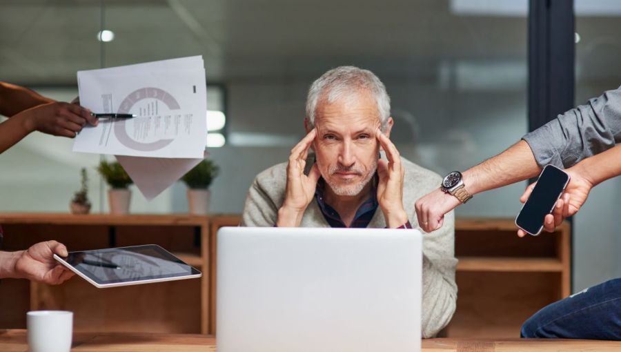 Gestresster Geschäftsmann im Büro vor Laptop, umgeben von Mitarbeitern mit Tablet, Smartphone und Dokumenten.
